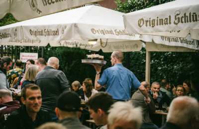Biergarten der Hausbrauerei Zum Schlüssel. Ein Kellner in blauem Hemd trägt ein Tablett mit mehreren frisch gezapften Gläsern Altbier durch einen belebten Außenbereich unter weißen Sonnenschirmen mit dem Aufdruck „Original Schlüssel“.