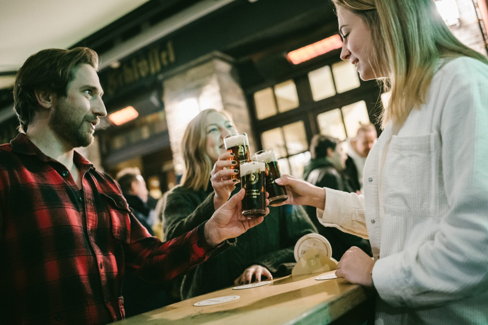 Drei Personen stoßen im Brauhaus „Zum Schlüssel“ an der Theke mit Gläsern dunklem Bier an und unterhalten sich in geselliger Atmosphäre.
